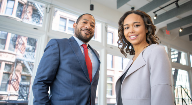 Two smiling people in business attire looking towards the camera. They are in a modern office building in a city.